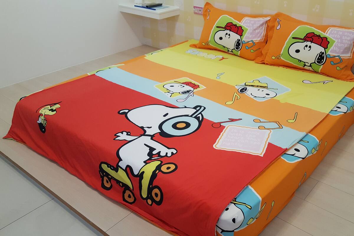 A bed with a Snoopy-themed bedspread and pillows, a small bedside table with a lamp, and a wall with a musical note pattern.