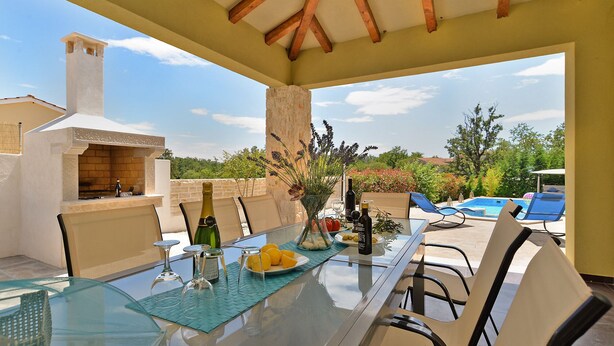 An outdoor dining area with a glass table, chairs, a pizza oven, and a view of a swimming pool and greenery. - Villa 1519/15239 (Istria - Žminj), 25000m from the beach (Žminj)