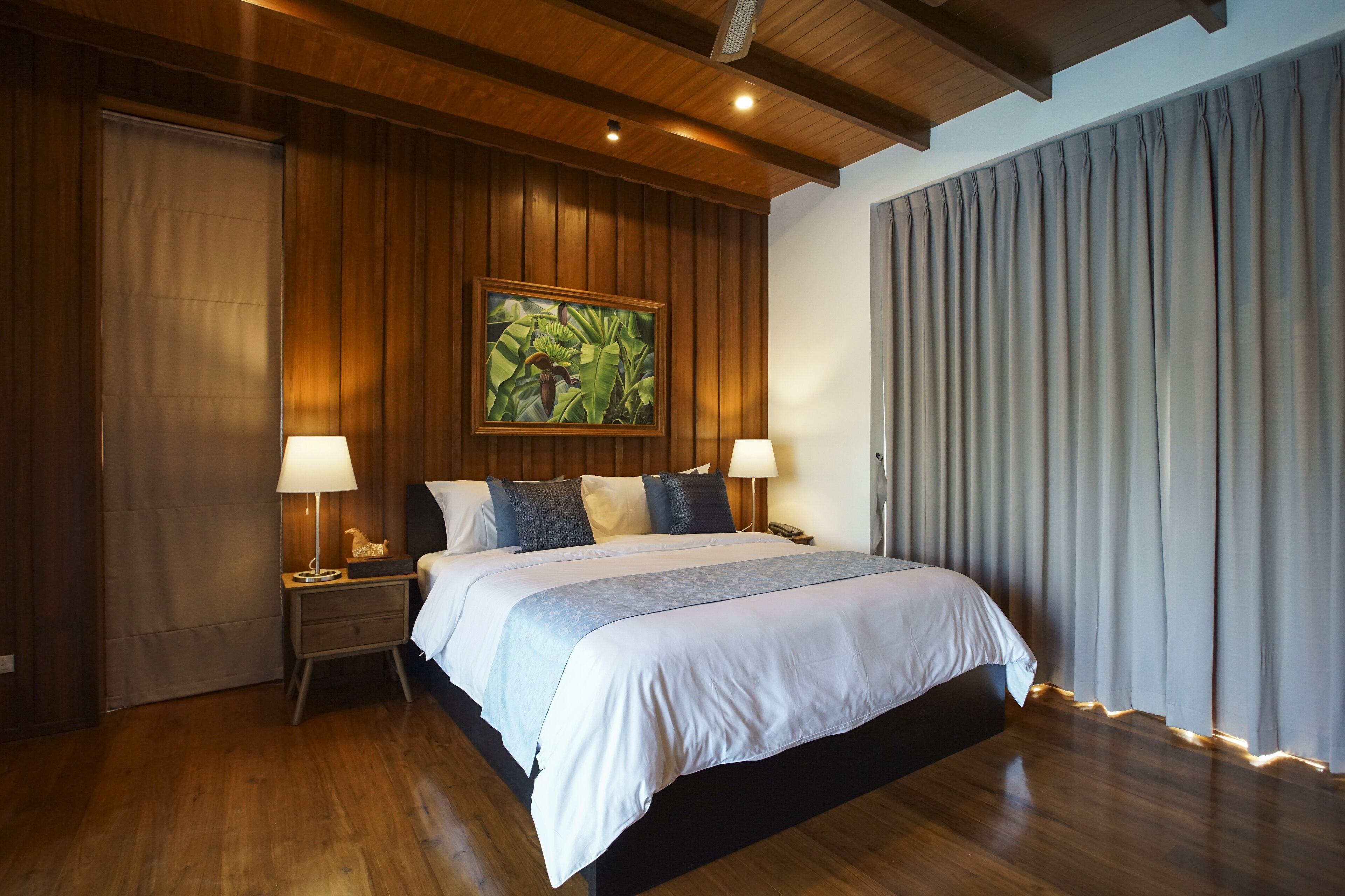 Un dormitorio con una cama grande, paredes de paneles de madera, un cuadro, dos lámparas de noche y una ventana con cortinas.