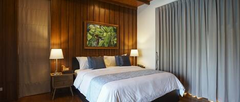 Un dormitorio con una cama grande, paredes de paneles de madera, un cuadro, dos lámparas de noche y una ventana con cortinas.