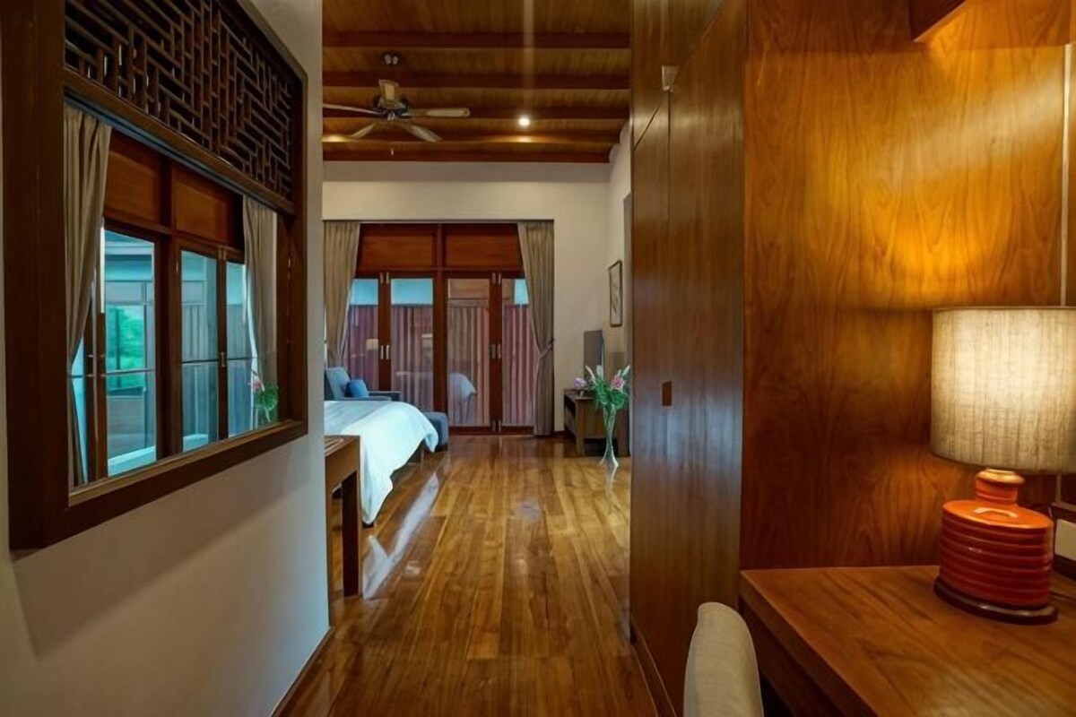 Quarto de hotel com piso de madeira, uma cama, uma escrivaninha com luminária e vista pela janela.