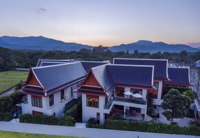 Una casa moderna con varios niveles y un balcón, rodeada de exuberante vegetación y montañas al fondo.
