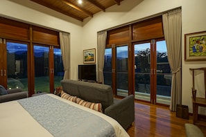 A bedroom with a large bed, a sofa, and a TV. There are large windows with curtains and a painting on the wall.
