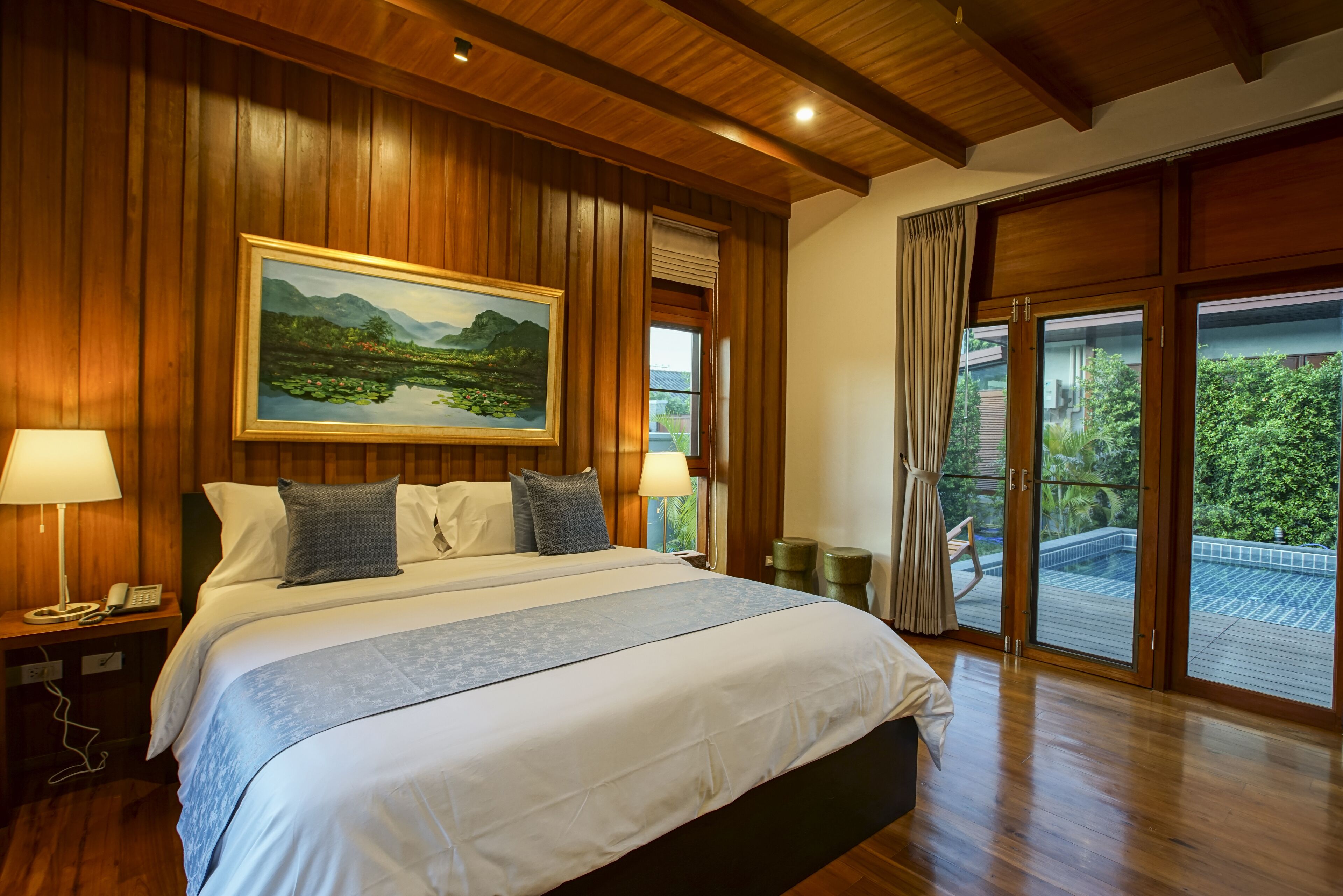 A bedroom with a large bed, wooden paneling, a painting of a landscape, a balcony with a view, and a telephone on the nightstand.