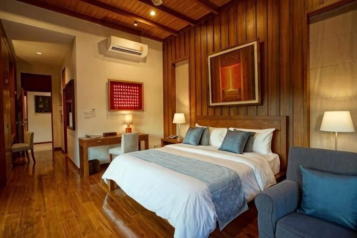 Habitación de hotel con techo de madera, una cama grande con colcha azul y blanca, dos mesitas de noche con lámparas, un escritorio con silla y un adorno rojo en la pared.