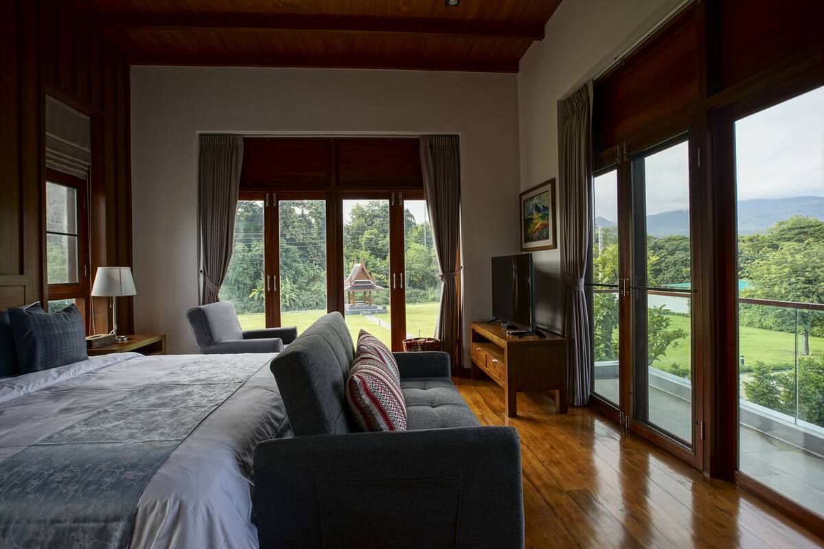 Un dormitorio amplio con una cama grande, un sofá y un televisor. Cuenta con ventanas grandes que ofrecen vistas a zonas verdes y montañas.