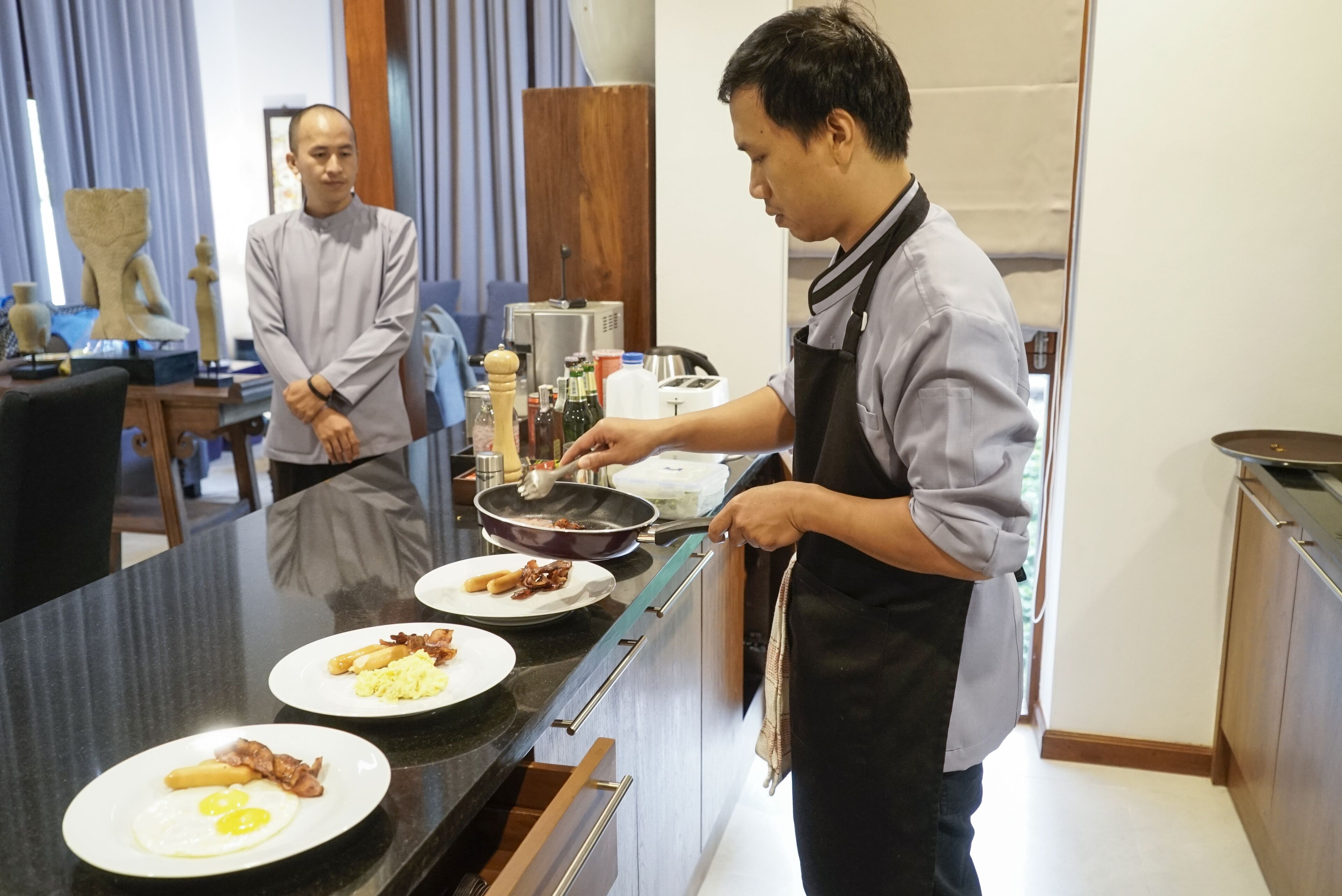 Una cucina con una persona che cucina, un tavolo da pranzo con piatti di cibo e una persona in piedi sullo sfondo.