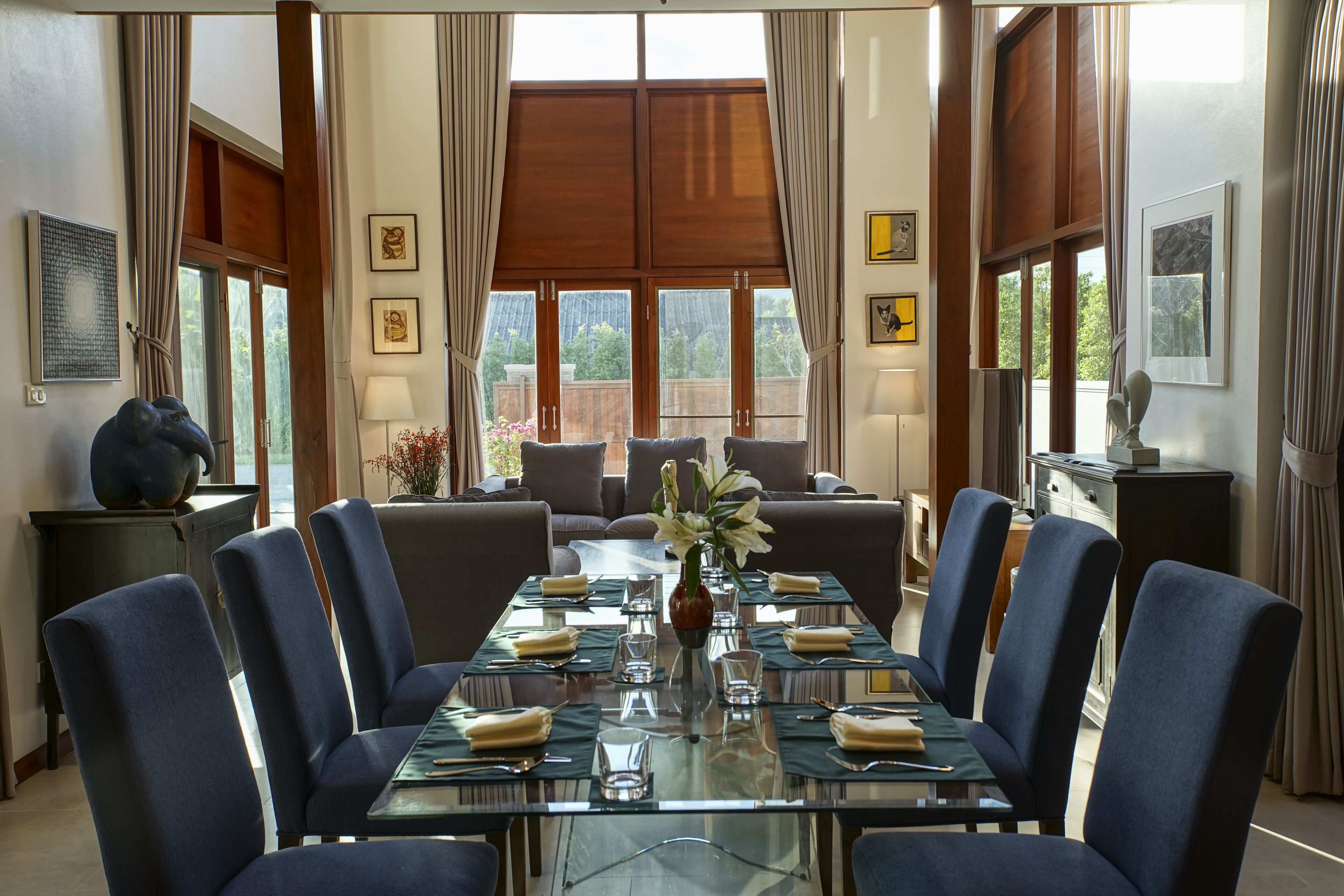 A dining area with a glass table set for six, surrounded by blue chairs, with a view of trees through large windows.