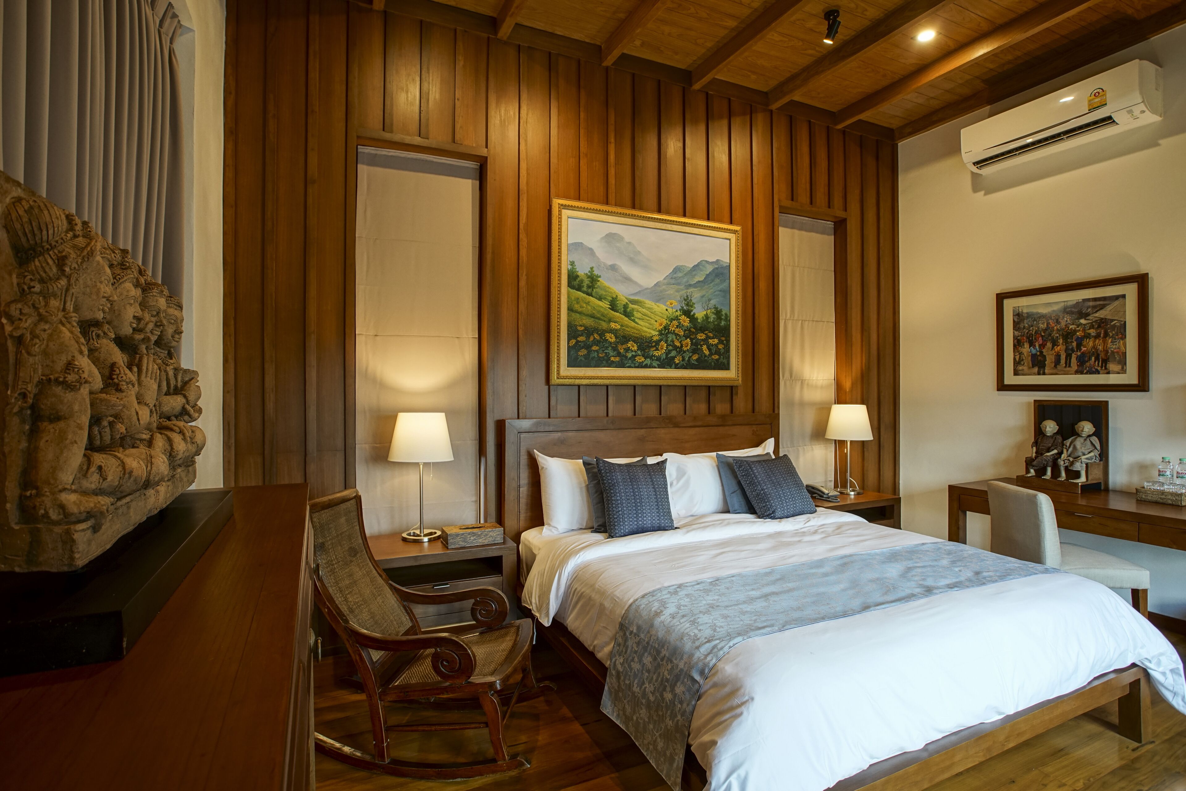 A hotel room with a wooden paneled wall, a bed with white linens, a painting of a landscape, a desk with a chair, and an air conditioning unit.