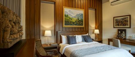 A hotel room with a wooden paneled wall, a bed with white linens, a painting of a landscape, a desk with a chair, and an air conditioning unit.