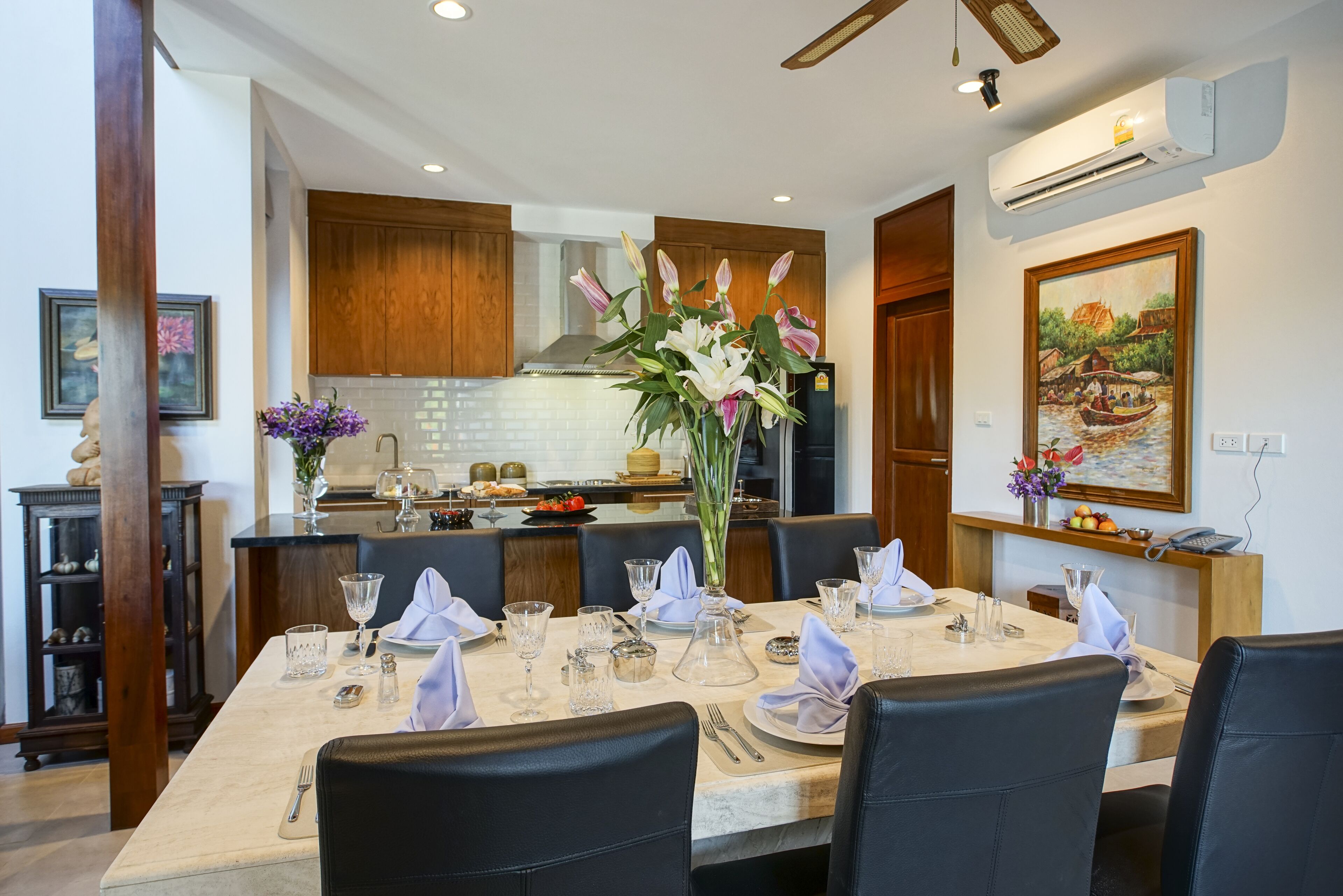 A dining area with a table set for four, a kitchen in the background, and a painting on the wall.