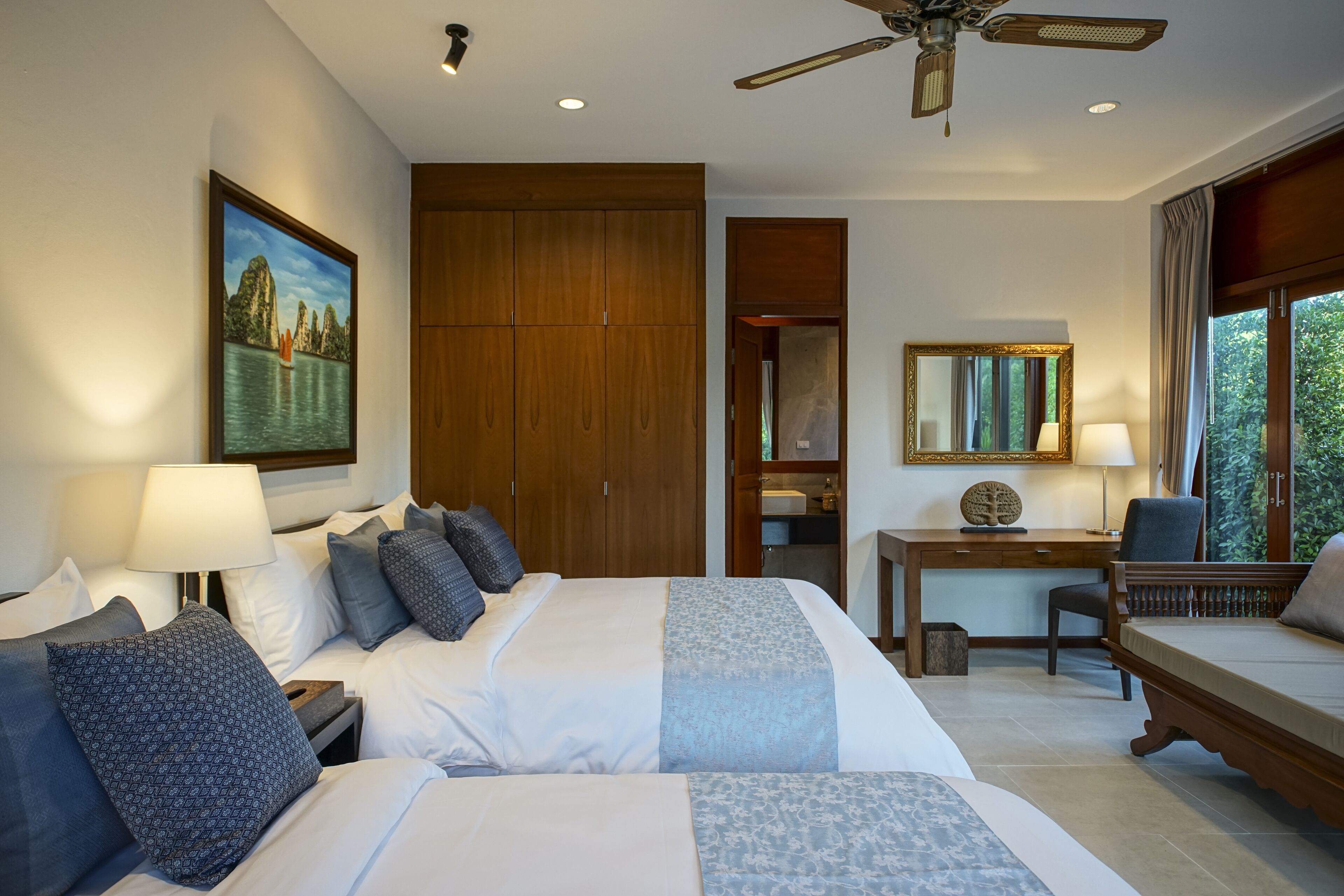 Ein Hotelzimmer mit zwei Betten, einem Schreibtisch, einem Sessel, einem Deckenventilator und einem großen Fenster.
