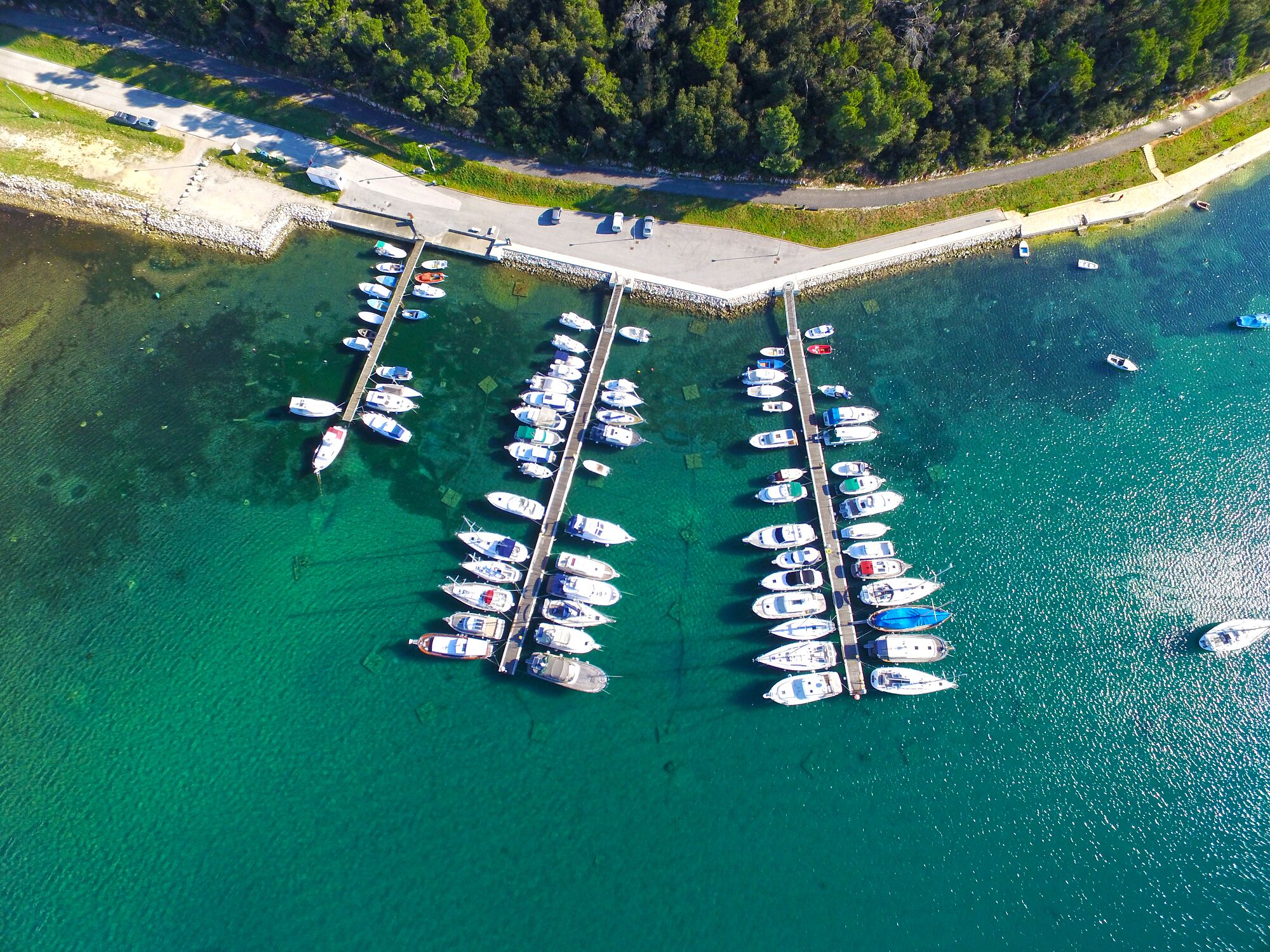 Vue aérienne d’une marina où de nombreux bateaux sont amarrés aux quais, entourée d’une eau turquoise cristalline et d’une zone boisée.