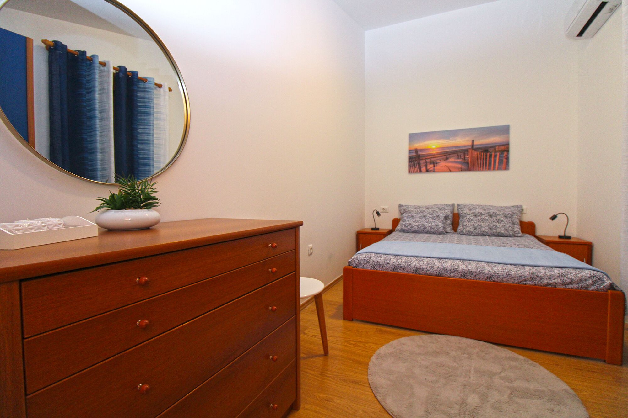 A bedroom with a wooden dresser, a bed with patterned bedding, a small potted plant, a round rug, and a painting on the wall.