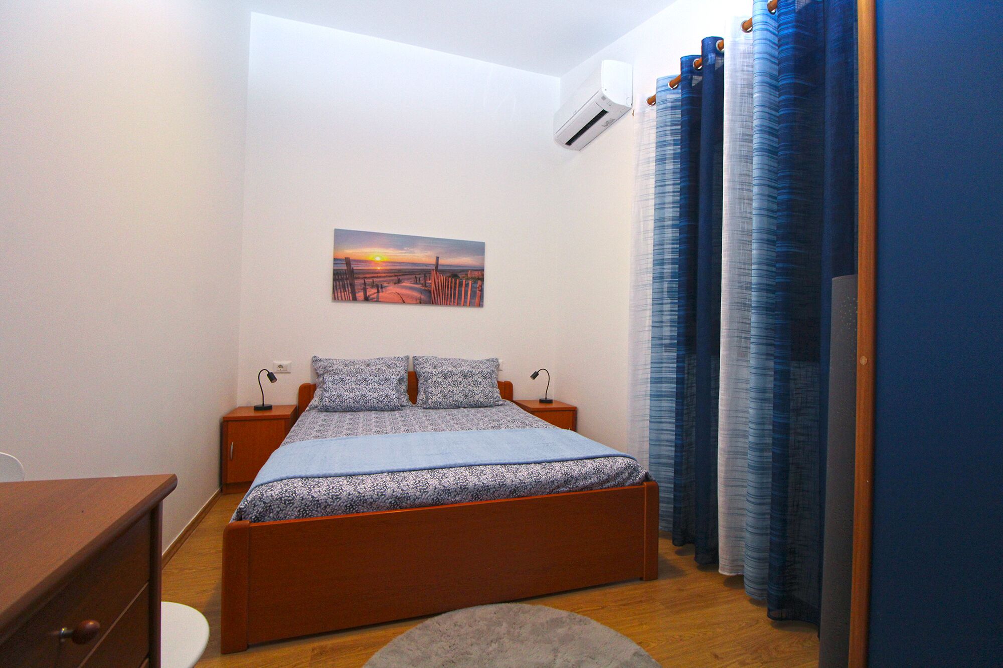 A bedroom with a bed, a nightstand, an air conditioner, a painting, and blue curtains.
