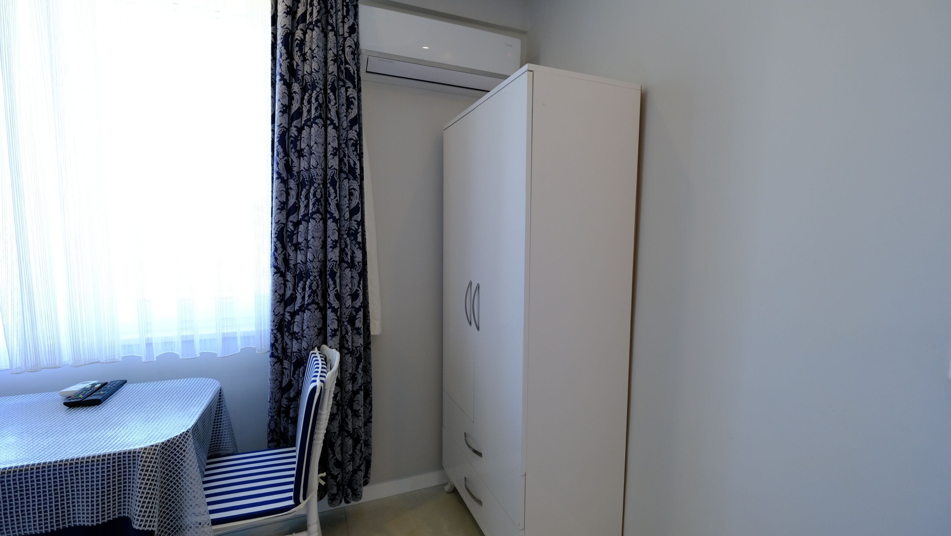 A small room with a white wardrobe, a table with a remote control, and a chair.