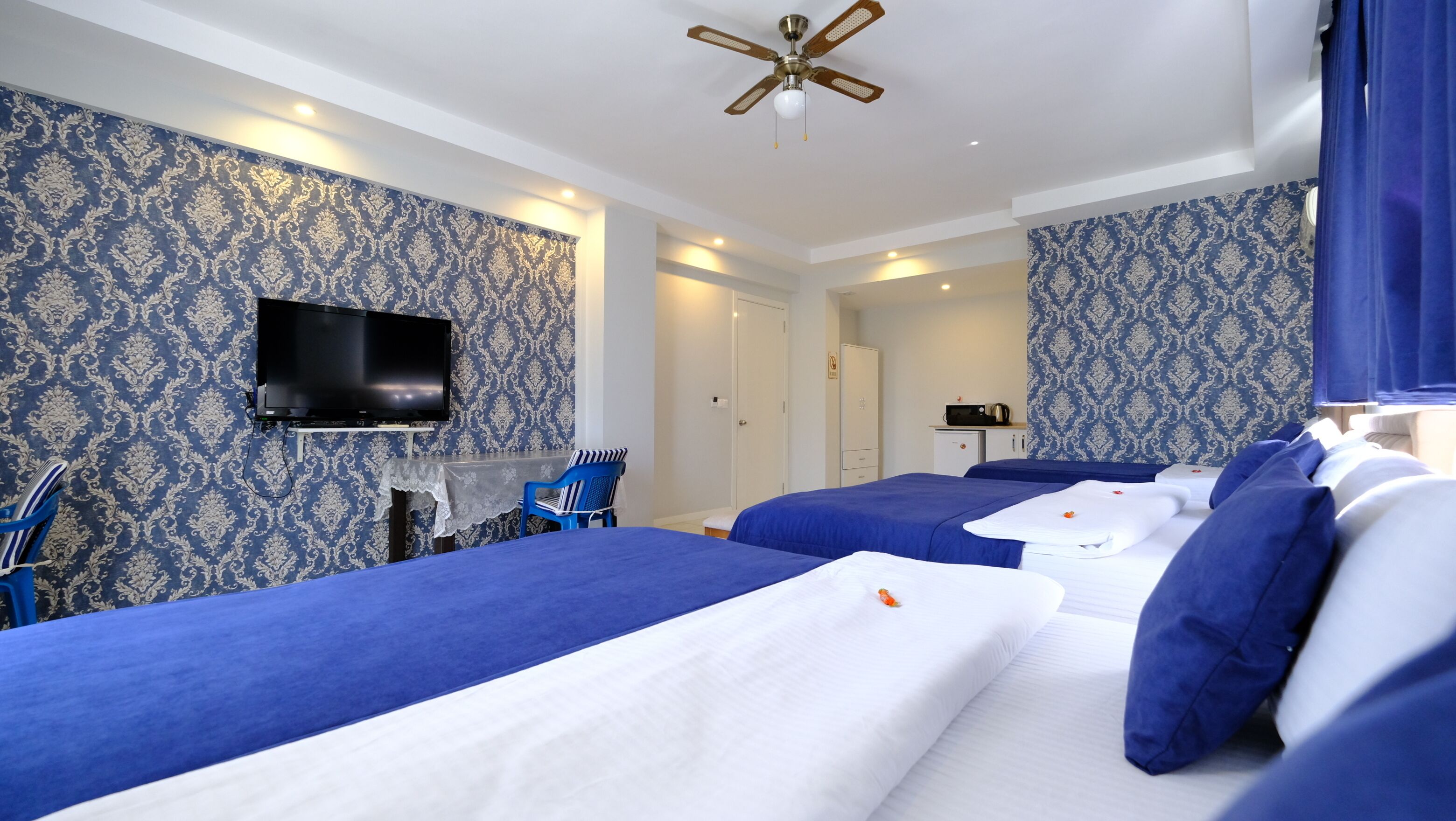 Ein Hotelzimmer mit zwei Betten, einem Fernseher, einem Deckenventilator und einer kleinen Küchenzeile.
