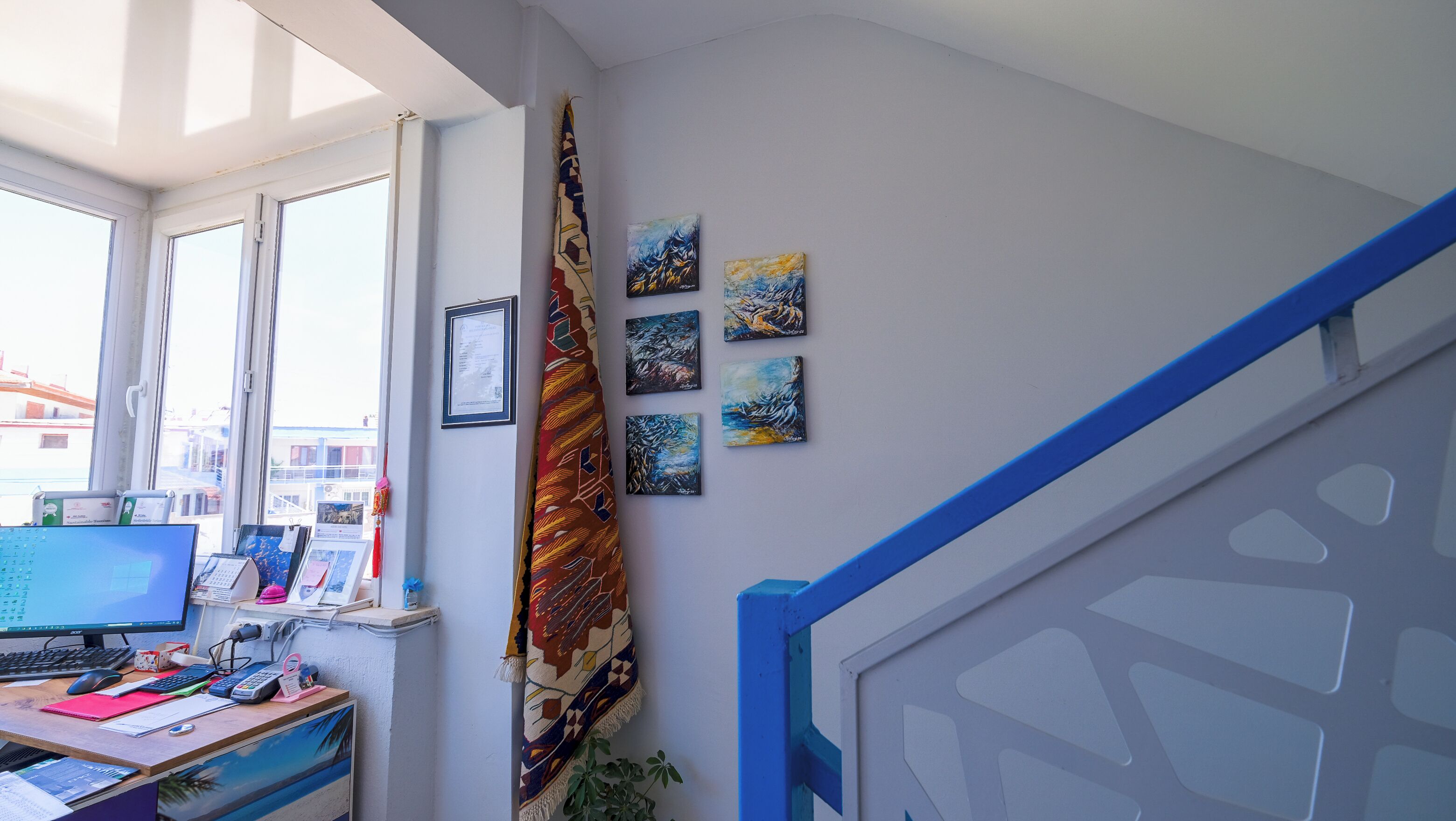 A room with a desk, computer, and a window with a view of buildings. There are paintings on the wall and a blue railing.