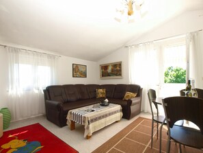 A living room with a brown sofa, a small table with a striped tablecloth, a red rug with a cartoon character, and a window with white curtains.