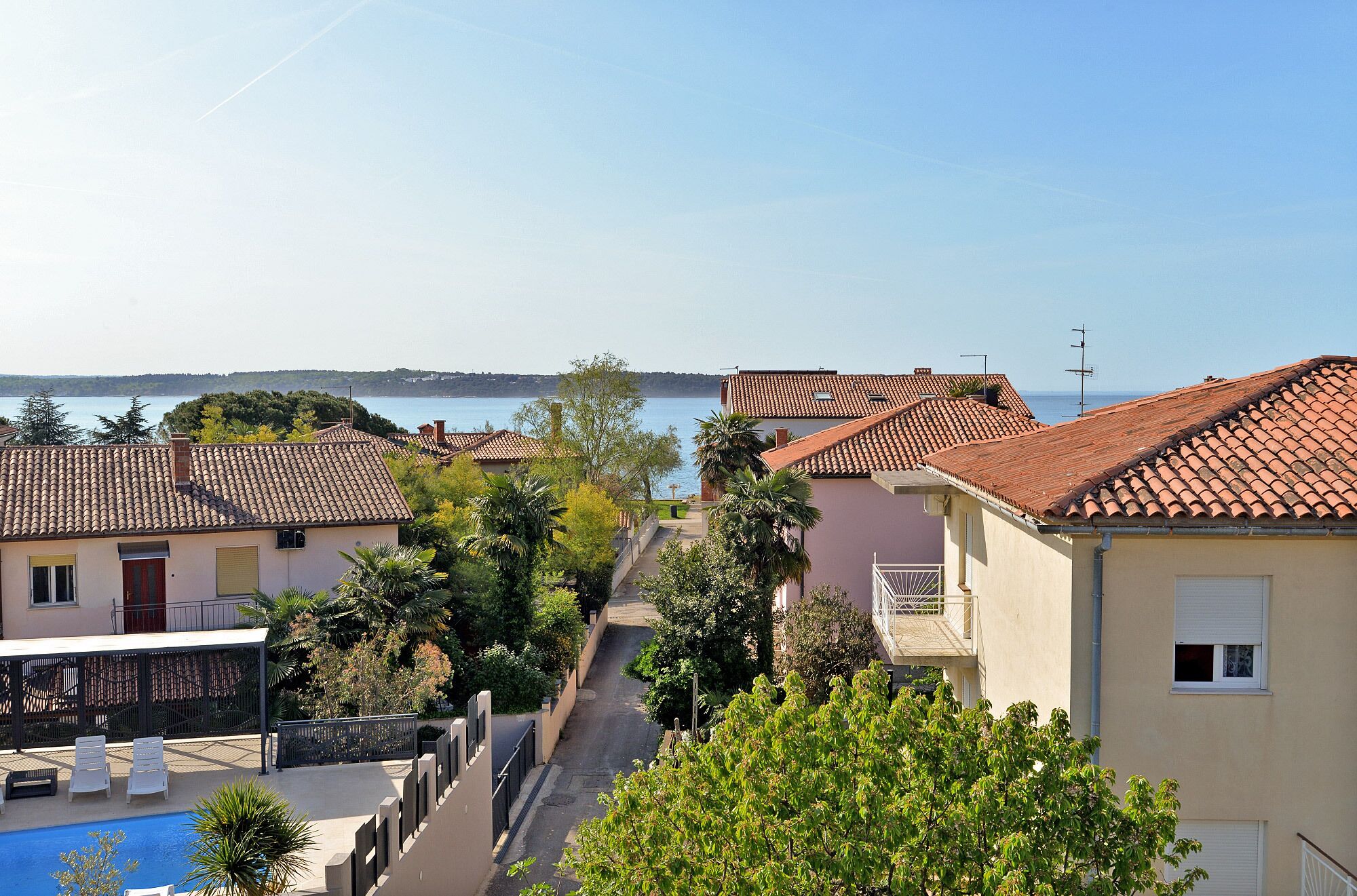 A coastal residential area with terracotta-tiled roofs, a swimming pool, and a clear view of the sea.