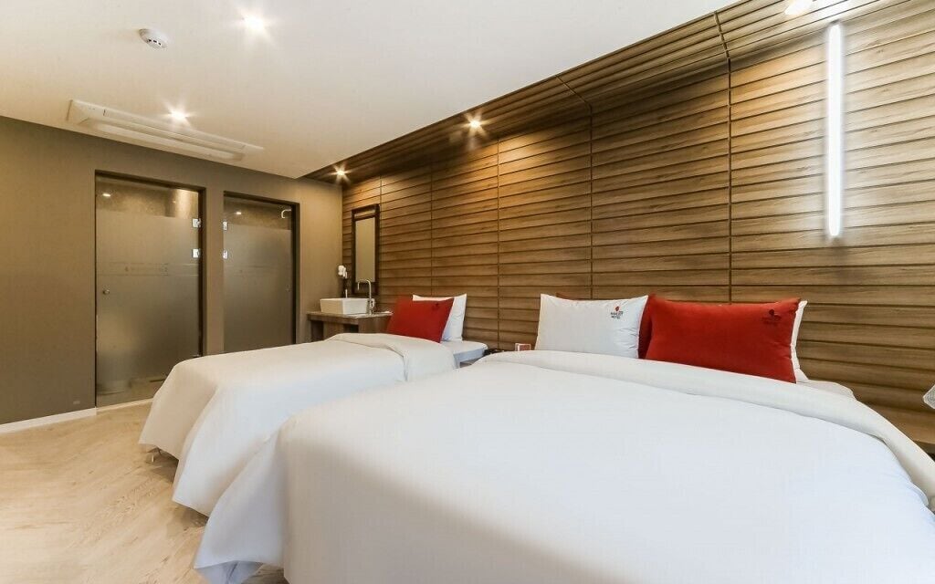 A modern hotel room with two beds, a wooden panel wall, a small bedside table, and a mirror.