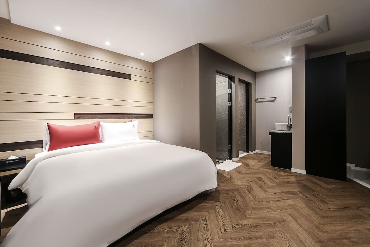 A modern hotel room with a large bed, a wooden headboard, a small bedside table, and a walk-in closet.