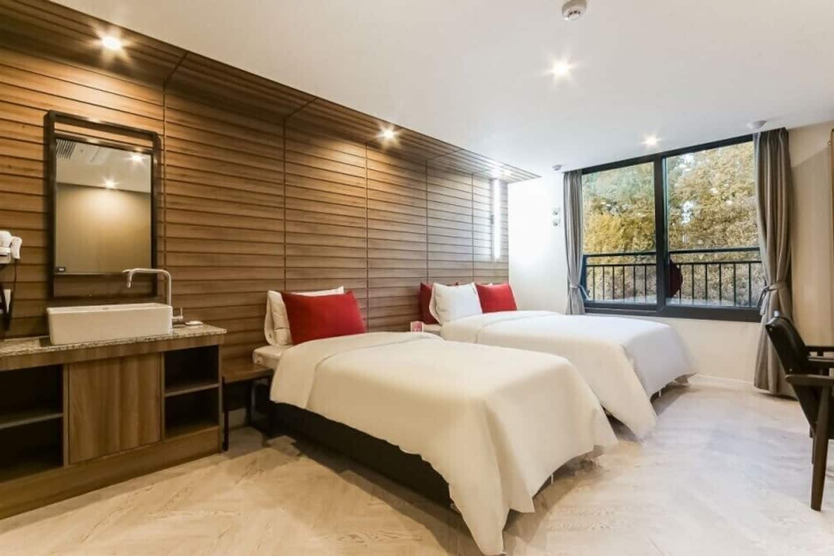 A modern hotel room with a wooden wall panel, a large window with a view, a sink, and two beds with white bedding and red pillows.
