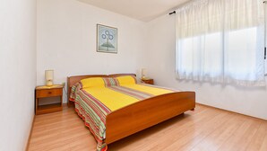 A bedroom with a wooden bed, two bedside tables with lamps, a window with sheer curtains, and a framed picture on the wall.