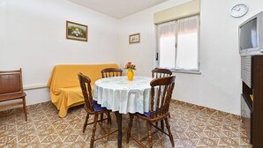 A dining area with a round table, four wooden chairs, a yellow sofa, a clock, and a television on a cabinet.