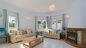 A spacious living room with two beige sofas, a wooden coffee table, a stone fireplace, and large windows with curtains. - Charlotte - Benissa, Costa Blanca (Benissa)