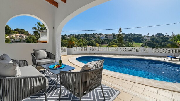 A poolside area with wicker furniture, a table with fruit, and a view of a hillside with houses. - Charlotte - Benissa, Costa Blanca (Benissa)
