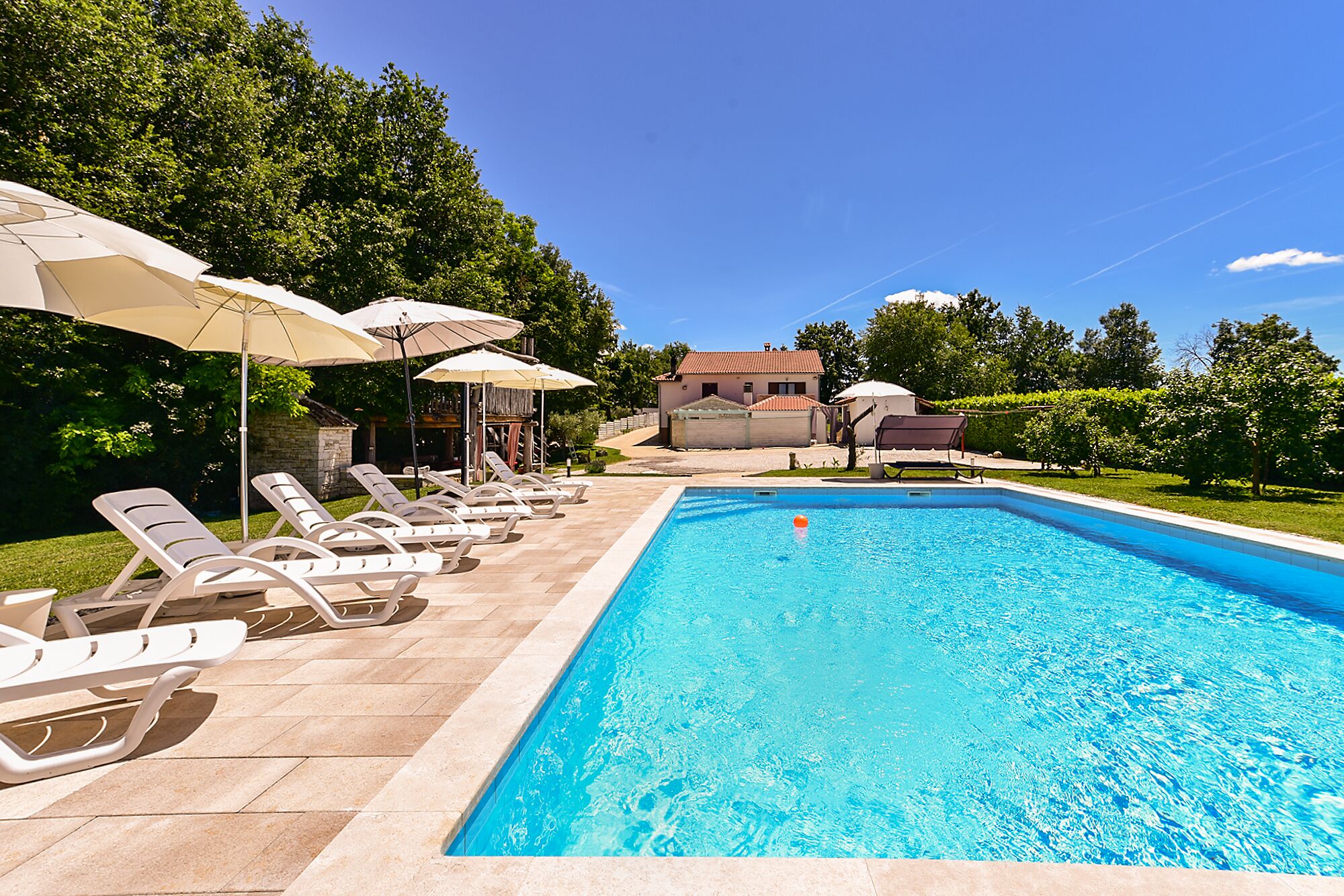 A swimming pool with lounge chairs and umbrellas, surrounded by greenery and a building in the background.