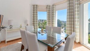 A modern dining area with a glass table and white chairs, large windows with striped curtains, and a view of a cityscape. - Modern and comfortable holiday home in Benissa, Costa Blanca, Spain  with private pool for 6 persons (Benissa)