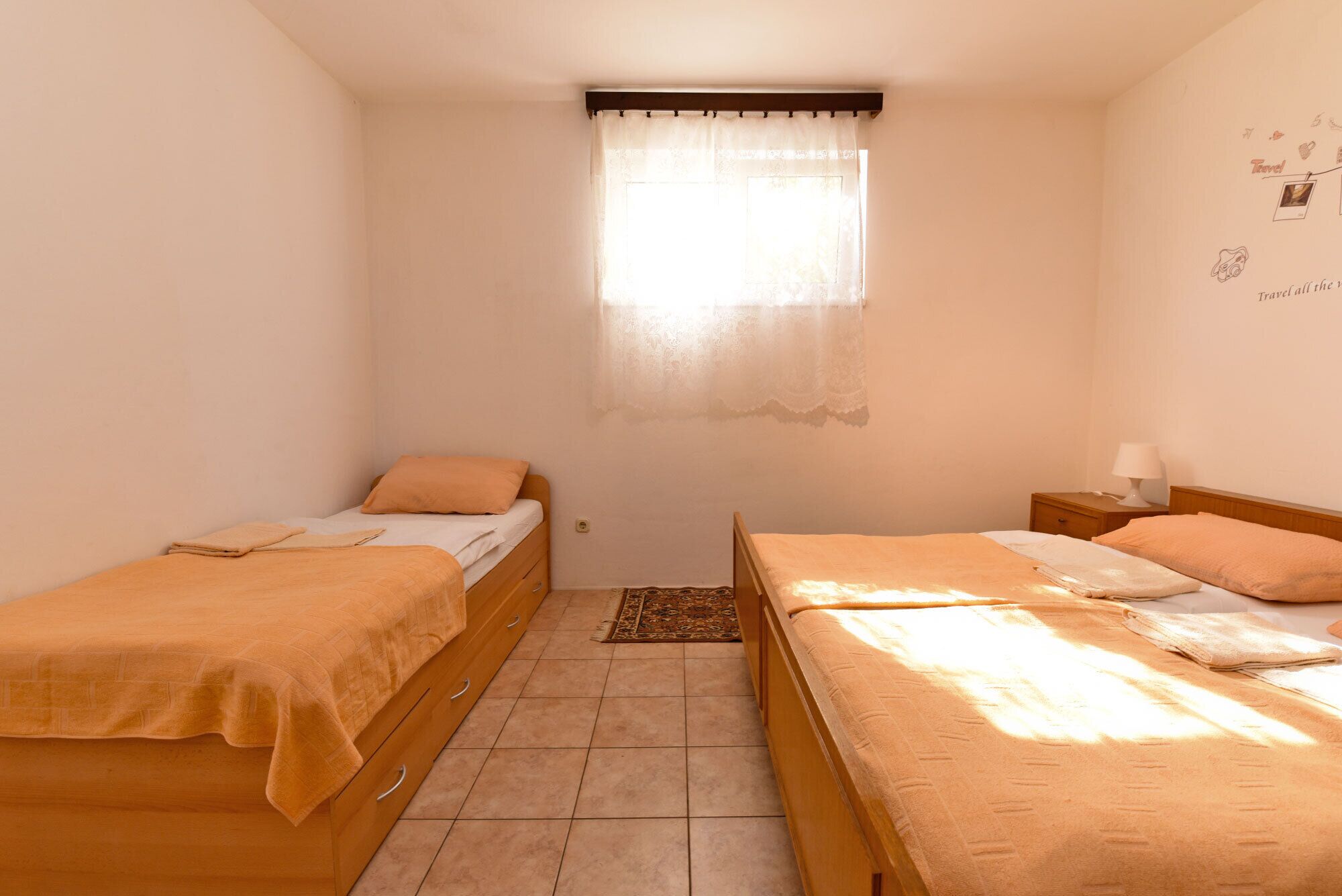 Quarto pequeno e bem iluminado com duas camas de solteiro, uma janela com cortinas leves, mesa de cabeceira com abajur e decoração temática de viagens nas paredes.
