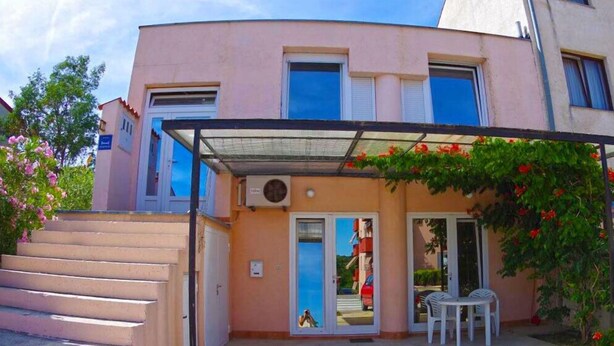 Ein zweistöckiges Gebäude mit flachem Dach, einer überdachten Eingangspforte mit Glastür sowie einer kleinen Außen-Sitzgelegenheit mit zwei Stühlen.