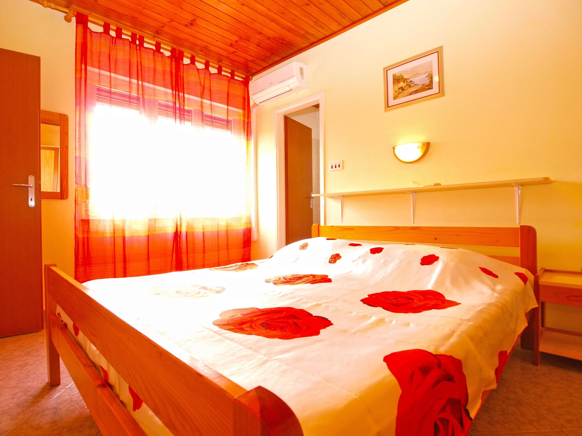 Une chambre à coucher avec un lit en bois, une fenêtre avec des rideaux rouges, un luminaire fixé au mur et une porte.
