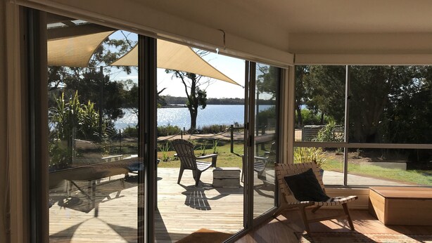 Habitación iluminada por el sol con amplios ventanales que dan a una terraza con mobiliario exterior y vistas a un lago.