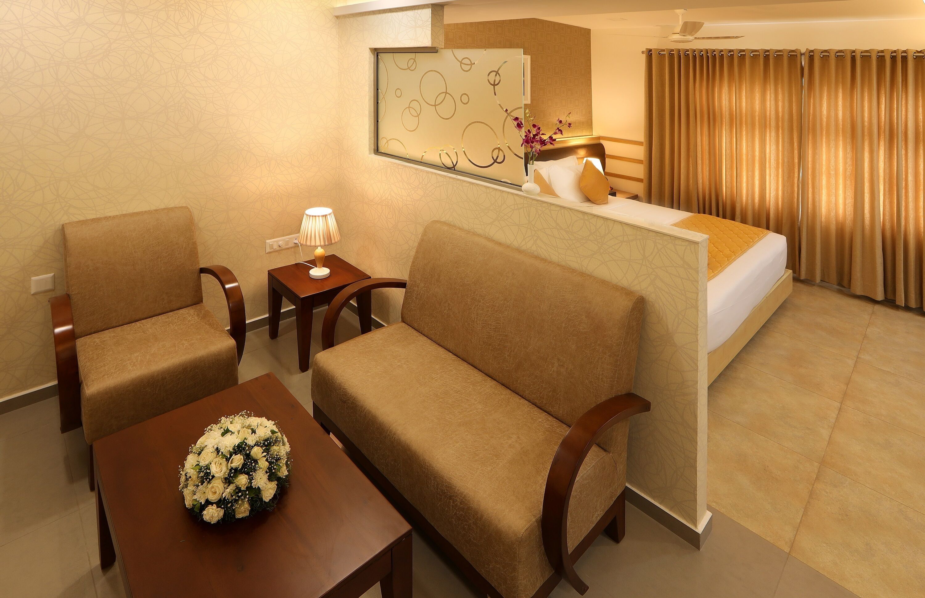 Habitación de hotel con sofá, una mesita con un arreglo floral, una mesita de noche con lámpara, una cama con cabecera y una ventana con cortinas.