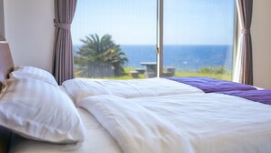 A bed with white and purple bedding, a window with curtains, and a view of the ocean. - Resort villa miko (Oshima)