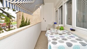 Un balcone con un tendalino a righe, un tavolo con una tovaglia decorata e una finestra che si affaccia su un edificio e auto parcheggiate.