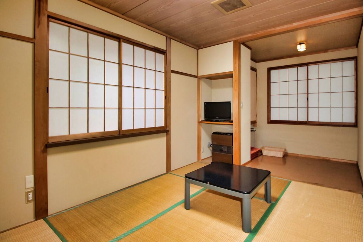 A traditional Japanese room with tatami flooring, a low table, a television on a shelf, and sliding doors.