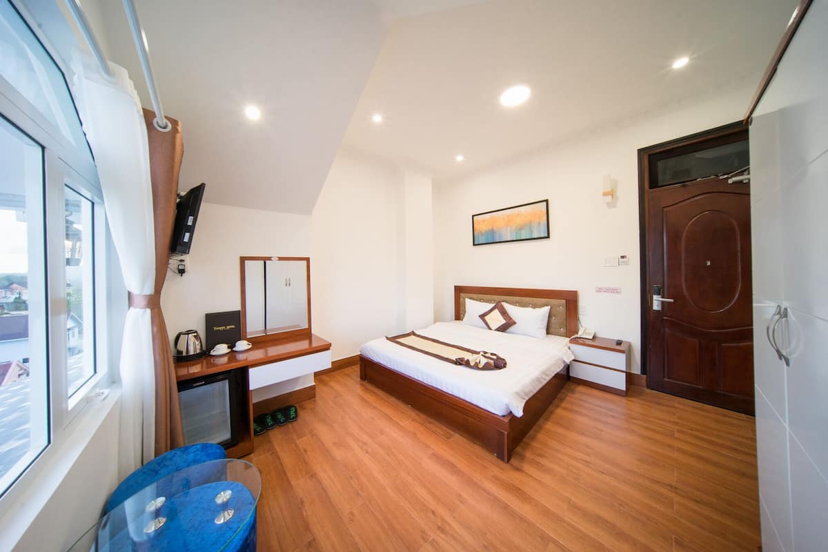 Habitación de hotel con piso de madera, cama con ropa blanca, escritorio con cafetera, espejo y vista al exterior.