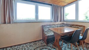 Een hoekzaak met een houten plafond, twee ramen met gordijnen, een bank met een bloemenkussen, een houten tafel en twee stoelen.