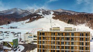 A modern building with a multi-story structure, a parking area with vehicles, and a ski slope in the background.