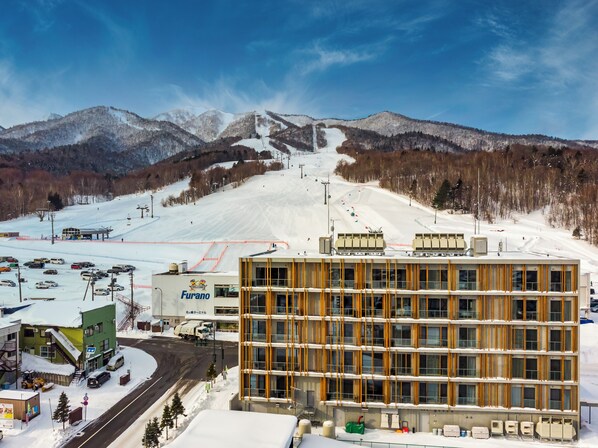 A modern building with a multi-story structure, a parking area with vehicles, and a ski slope in the background. - Fenix Furano (Furano)