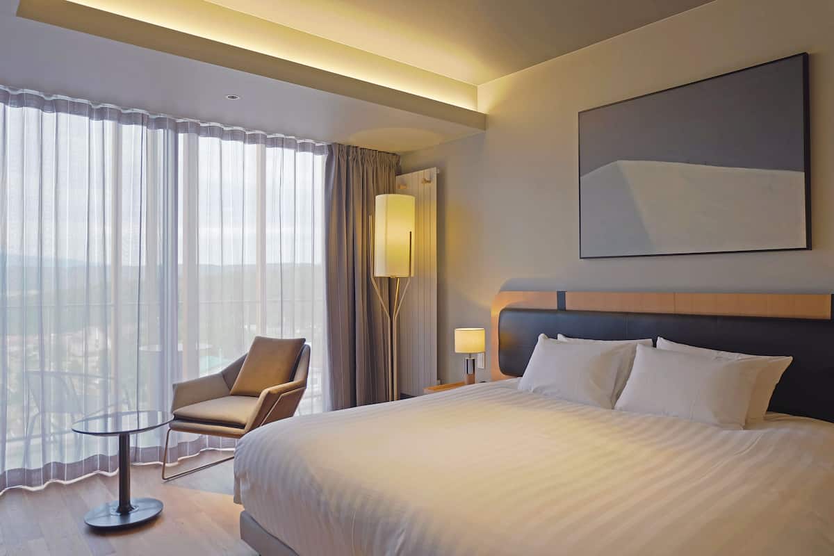 A hotel room with a large bed, a chair, a table, and a view of the outdoors.