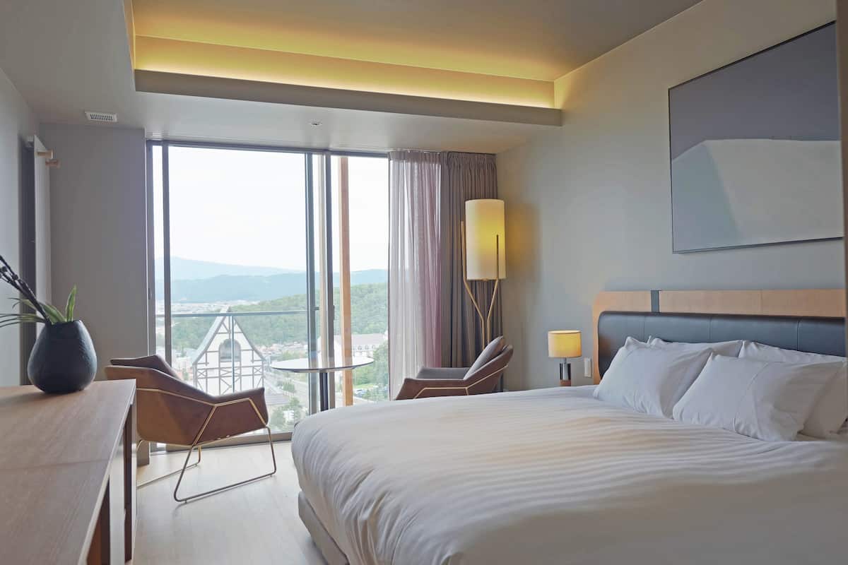A hotel room with a large bed, a desk, a chair, a lamp, and a view of the outdoors.
