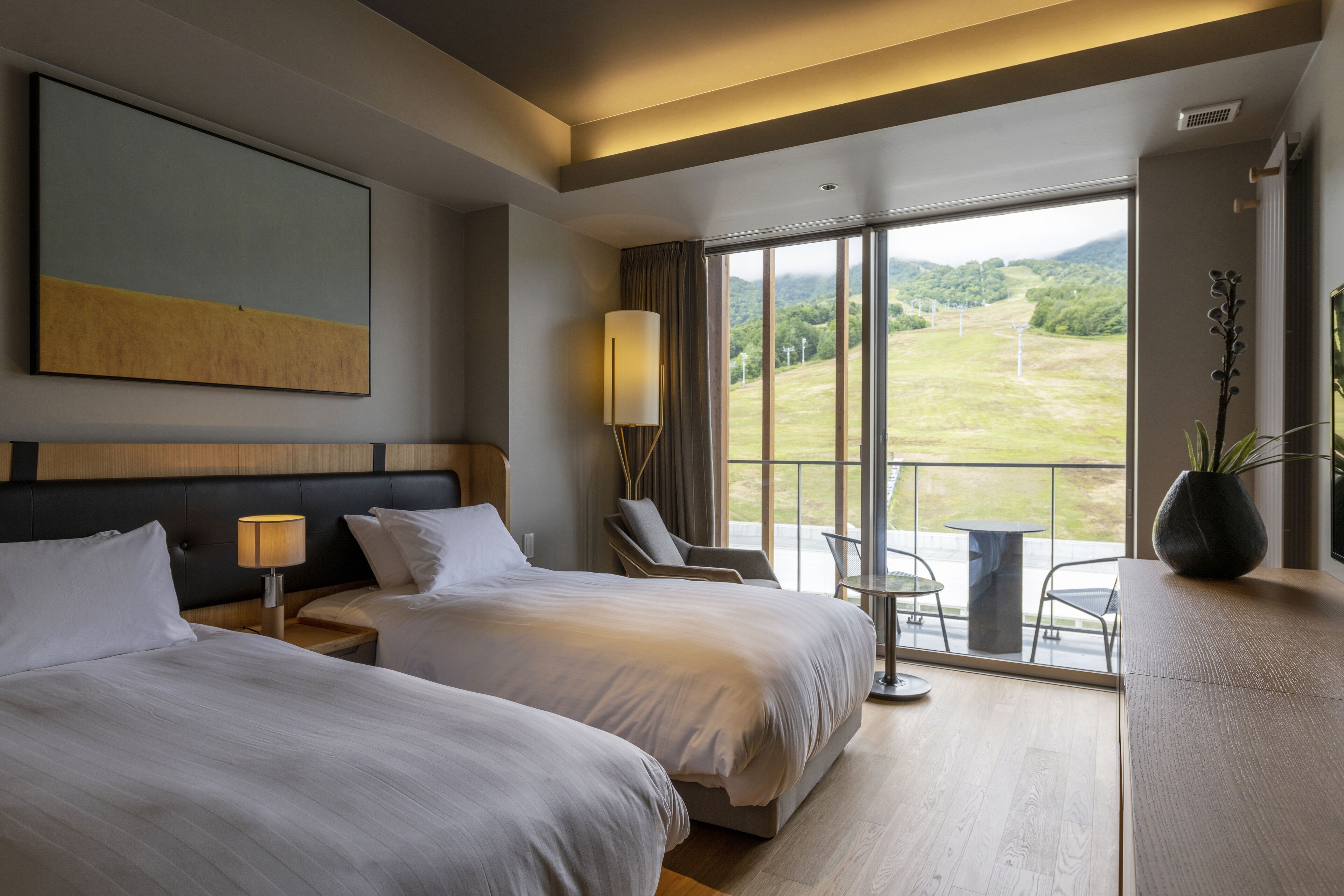 Habitación de hotel con dos camas, un ventanal con vistas a un paisaje verde, una mesita y una silla.