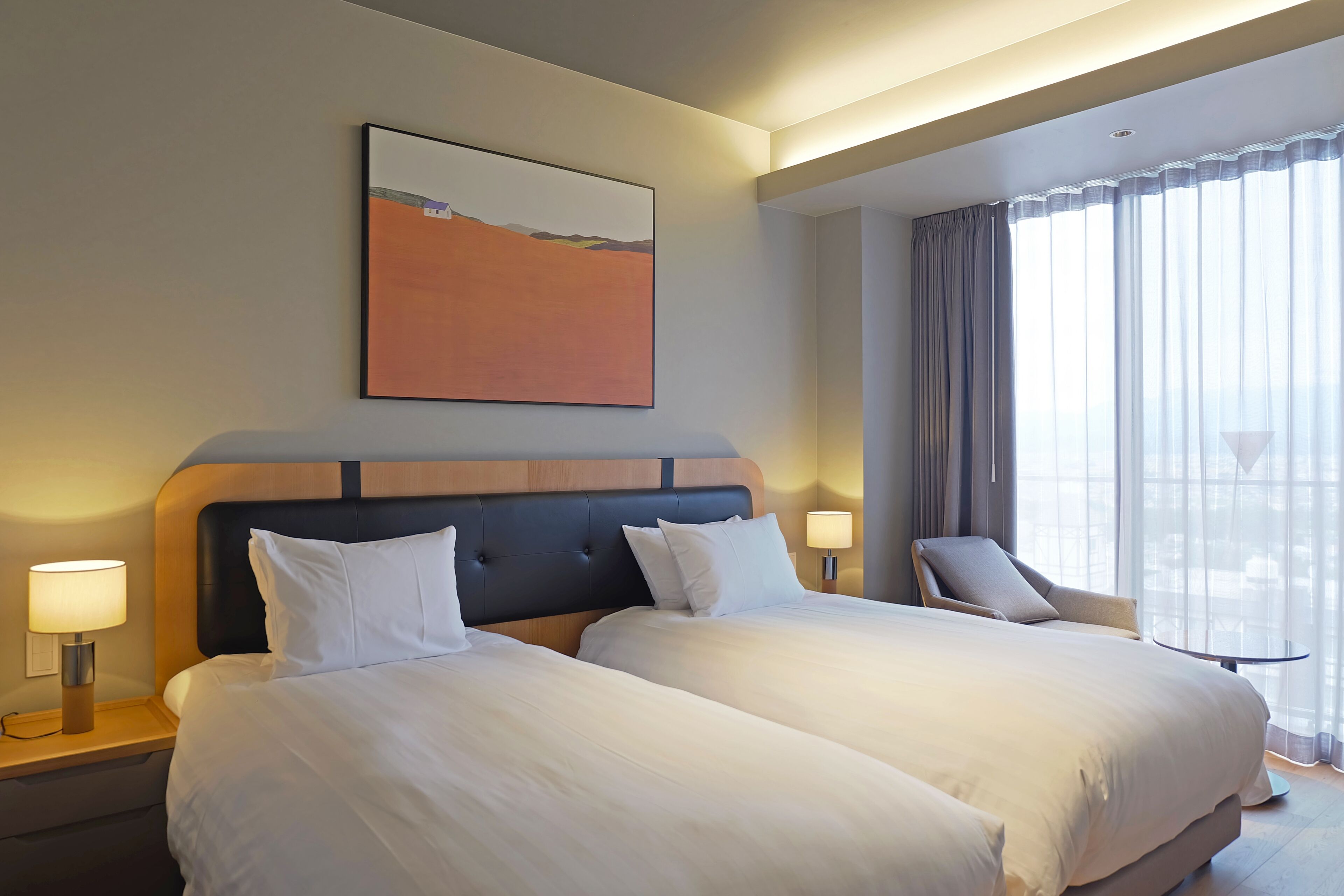 Habitación de hotel con dos camas, cabecero, un cuadro, una silla, una mesa y una ventana con cortinas.