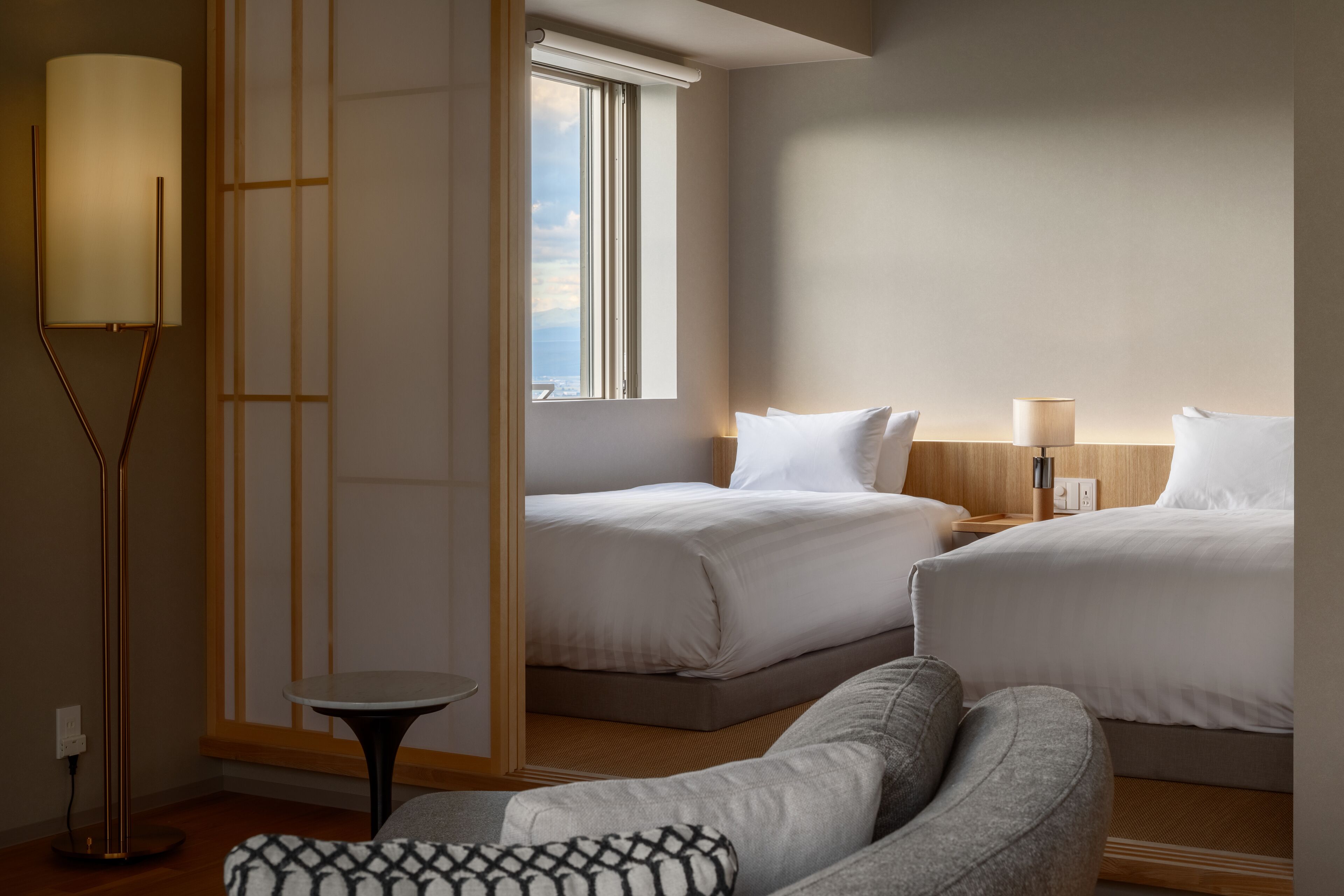 A hotel room with two beds, a standing lamp, a small table, and a view of the city through a window.